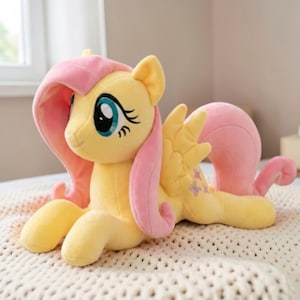 May include: A plush toy pony in a lying position. The pony is primarily yellow with pink accents on its mane, tail, and ears. It has large blue eyes and a pair of yellow wings with purple butterfly details. The toy is soft and cuddly.