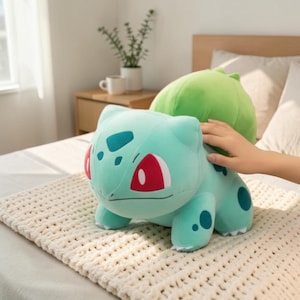 Op de afbeelding: Een lichtgroen Bulbasaur-knuffel met rode ogen en turquoise vlekken. Het speelgoed heeft een grote groene bol op de rug. Het speelgoed ligt op een crèmekleurige, getextureerde deken.