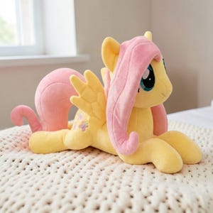 May include: A plush toy of a yellow pony with pink hair and wings, lying on a white knitted blanket. The pony has blue eyes and a small flower design on its side. The toy is soft and cuddly.