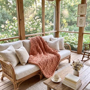 May include: A cozy outdoor living space with a rattan sofa, cream cushions, and a chunky knit orange blanket. A wooden coffee table holds books and a teacup. The space is surrounded by windows overlooking a lush garden.