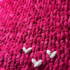 May include: A vibrant pink, chunky knit blanket with three white heart appliqu&eacute;s. The blanket has a plush, textured appearance, perfect for adding warmth and a pop of color to any space. The hearts are evenly spaced.
