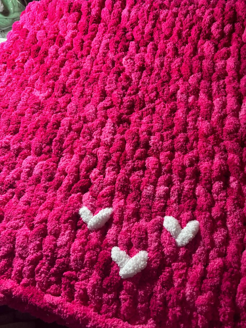 May include: A vibrant pink, chunky knit blanket with three white heart appliqu&eacute;s. The blanket has a plush, textured appearance, perfect for adding warmth and a pop of color to any space. The hearts are evenly spaced.