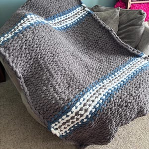 Puede incluir: Una manta de punto grueso en gris con un dise&ntilde;o a rayas en blanco, azul y gris. La manta est&aacute; hecha de hilo grueso y suave y est&aacute; colocada sobre un sof&aacute; gris. Esta acogedora manta es perfecta para acurrucarse.