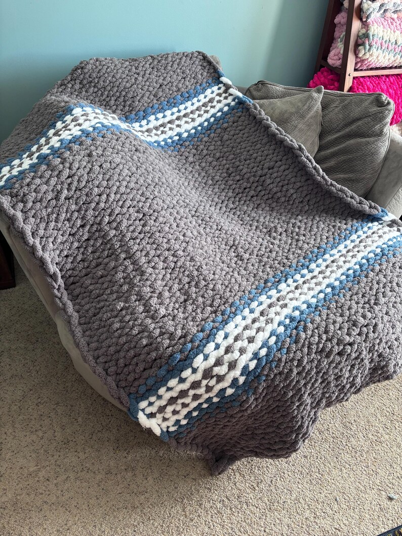 Puede incluir: Una manta de punto grueso en gris con un dise&ntilde;o a rayas en blanco, azul y gris. La manta est&aacute; hecha de hilo grueso y suave y est&aacute; colocada sobre un sof&aacute; gris. Esta acogedora manta es perfecta para acurrucarse.