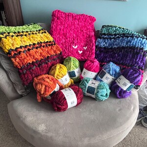May include: A collection of colorful, chunky knit blankets and yarn skeins. The blankets feature vibrant rainbow, pink, and blue-purple gradients. Several skeins of yarn in various colors, including yellow, green, and pink, are also visible.