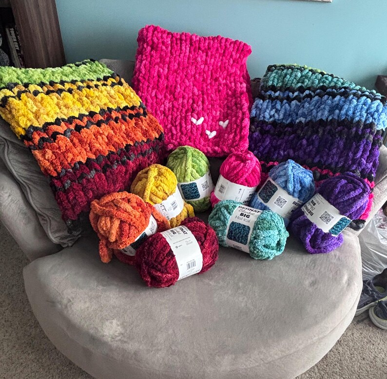 May include: A collection of colorful, chunky knit blankets and yarn skeins. The blankets feature vibrant rainbow, pink, and blue-purple gradients. Several skeins of yarn in various colors, including yellow, green, and pink, are also visible.
