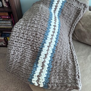Puede incluir: Una manta de punto grueso en tonos grises, con una franja central blanca, azul claro y gris. La manta est&aacute; sobre un sof&aacute;, mostrando su superficie texturizada y su dise&ntilde;o acogedor.