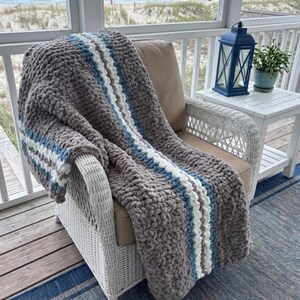 Puede incluir: Una manta de punto grueso gris con rayas blancas y azules, sobre una silla de mimbre blanca. La manta es gruesa y acogedora, ideal para acurrucarse en un porche con vistas al mar.