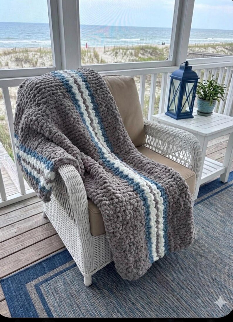 Puede incluir: Una manta de punto grueso gris con rayas blancas y azules, sobre una silla de mimbre blanca. La manta es gruesa y acogedora, ideal para acurrucarse en un porche con vistas al mar.