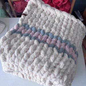 May include: A cream-colored, chunky knit blanket with a diagonal stripe pattern in light blue and pink. The blanket is made of a soft, plush material, perfect for snuggling. The texture is visible, with large, looped stitches.