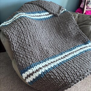Puede incluir: Una manta de punto grueso en color gris neutro, con rayas blancas y azul claro. La manta est&aacute; colocada sobre una silla, mostrando su textura y dise&ntilde;o. Esta acogedora manta es perfecta para a&ntilde;adir calidez y estilo a cualquier espacio habitable.