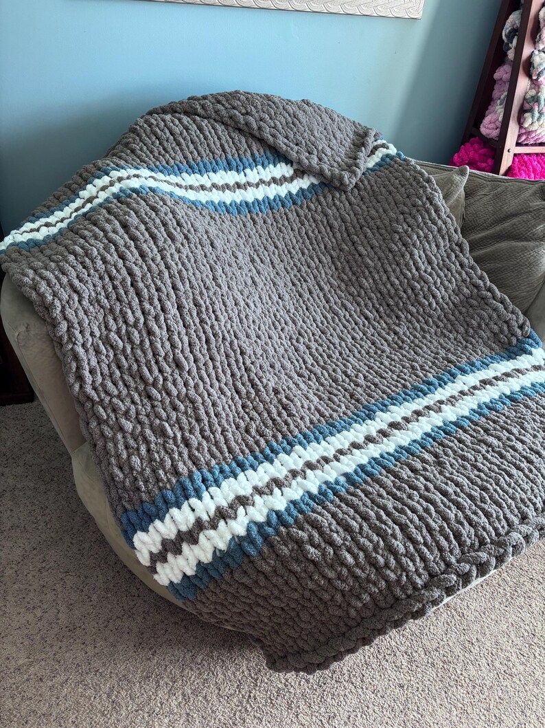 Puede incluir: Una manta de punto grueso en color gris neutro, con rayas blancas y azul claro. La manta est&aacute; colocada sobre una silla, mostrando su textura y dise&ntilde;o. Esta acogedora manta es perfecta para a&ntilde;adir calidez y estilo a cualquier espacio habitable.
