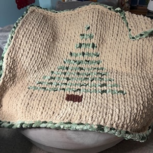 May include: A chunky knit blanket made of thick beige yarn featuring a central Christmas tree design created with variegated green yarn and a small brown rectangular trunk. The blanket has a decorative border of green and beige yarn.