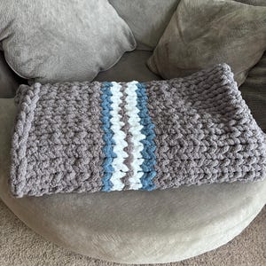Puede incluir: Una manta de punto grueso en tonos grises, con una franja central blanca y azul claro. La manta est&aacute; doblada y colocada sobre una superficie redondeada y tapizada, ideal para la decoraci&oacute;n del hogar.