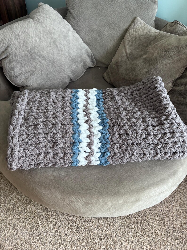 Puede incluir: Una manta de punto grueso en tonos grises, con una franja central blanca y azul claro. La manta est&aacute; doblada y colocada sobre una superficie redondeada y tapizada, ideal para la decoraci&oacute;n del hogar.