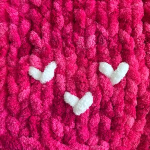 May include: Close-up of a handmade, chunky knit blanket in vibrant shades of pink. The blanket features three white heart-shaped appliqu&eacute;s, creating a playful design. The texture is soft and plush, perfect for a cozy home decor item.