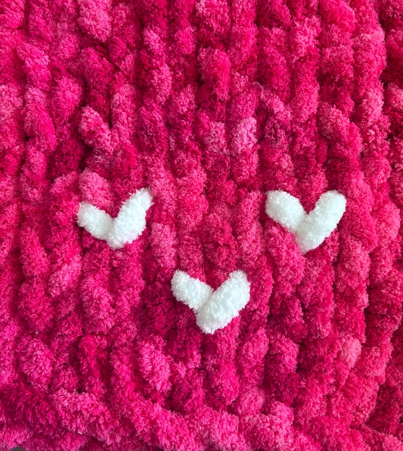 May include: Close-up of a handmade, chunky knit blanket in vibrant shades of pink. The blanket features three white heart-shaped appliqu&eacute;s, creating a playful design. The texture is soft and plush, perfect for a cozy home decor item.