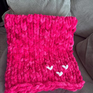 May include: A vibrant pink, chunky knit blanket with three white heart appliqu&eacute;s. The blanket is folded and resting on a neutral-colored sofa. The plush texture and bright color make it a cozy home decor item.
