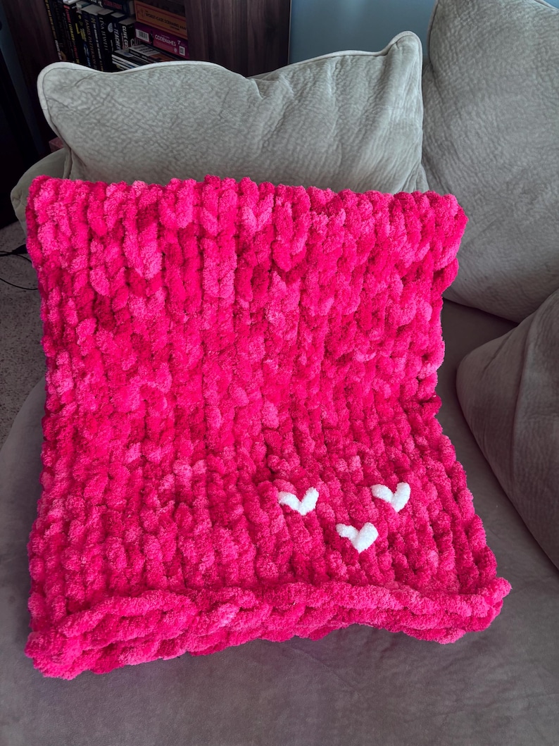 May include: A vibrant pink, chunky knit blanket with three white heart appliqu&eacute;s. The blanket is folded and resting on a neutral-colored sofa. The plush texture and bright color make it a cozy home decor item.