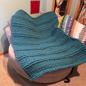 May include: A chunky knit blanket in a deep teal color featuring a textured chevron pattern with raised horizontal ridges. The blanket is draped over a round, light-colored upholstered chair, showcasing the thick, soft yarn loops throughout.