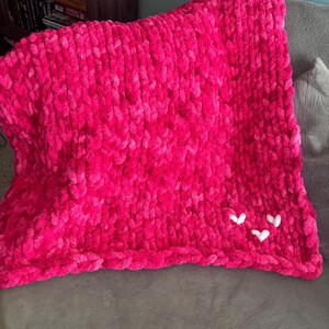May include: A vibrant pink, chunky knit blanket with three small white heart appliqu&eacute;s. The blanket is made of a soft, plush material and is folded over a gray surface. The blanket is a cozy home decor item.