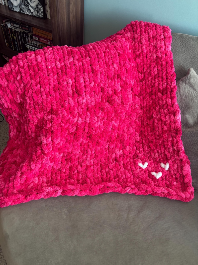 May include: A vibrant pink, chunky knit blanket with three small white heart appliqu&eacute;s. The blanket is made of a soft, plush material and is folded over a gray surface. The blanket is a cozy home decor item.