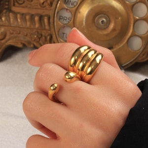 May include: A gold-colored ring set. The ring on the index finger has a small, rounded design. The ring on the middle finger has a thick, ribbed design with a rounded accent. The jewelry is shiny and reflective.