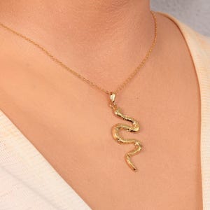 May include: A gold-coloured necklace with a snake pendant. The pendant has a textured surface and hangs from a fine gold chain. The necklace is displayed against a neutral background. The snake pendant is a unique design.