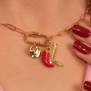 May include: Gold-toned necklace with a carabiner clasp and three charms: a cowboy hat, a red chili pepper, and a cowboy boot. The necklace is worn around the neck. The charms are detailed and add a touch of Western flair.