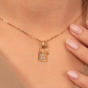 May include: A gold-colored necklace featuring a padlock pendant with the letter "A" encrusted with small, clear stones. A small key charm hangs beside the padlock. The necklace is on a delicate chain.