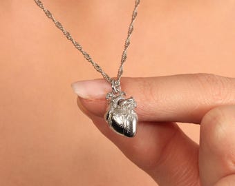 May include: A silver-colored necklace featuring a pendant shaped like a human heart. The heart pendant is intricately detailed and suspended from a delicate chain. The necklace is held by a person, showcasing the jewelry against a neutral background.