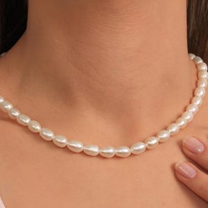 May include: A delicate pearl necklace with oval-shaped, off-white pearls. The necklace is draped around a person's neck, with a hand gently touching the pearls. The pearls have a subtle luster, reflecting the light.