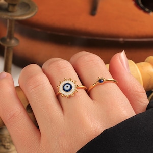 May include: Two gold rings are shown on a hand. One ring has a white and blue evil eye design with a gold beaded border. The other ring is a thin gold band with a small blue gemstone. The rings are on a person's finger.