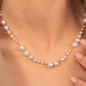 Può includere: Una delicata collana d'oro con perle bianche di varie dimensioni. Le perle sono disposte in modo graduato, creando un effetto visivo accattivante. La collana è presentata su uno sfondo neutro, evidenziando il suo design elegante.
