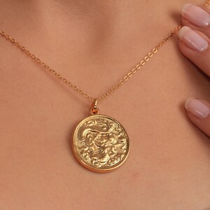 May include: A gold-coloured necklace with a round pendant featuring a detailed relief design. The pendant hangs from a delicate chain. The necklace is displayed on a person's neck.