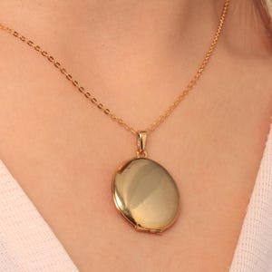 Könnte beinhalten: Ein goldfarbener, ovaler Medaillon-Anhänger an einer feinen Kette. Das Medaillon hat eine glatte, glänzende Oberfläche, die das Licht reflektiert. Kette und Anhänger haben einen passenden Goldton und bilden ein klassisches Schmuckstück.