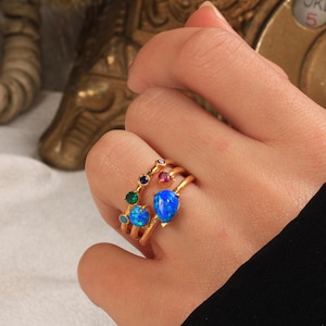 May include: Gold rings with colorful gemstones. One ring features a series of small, round gems in various colors. Another ring has two blue teardrop-shaped opals. The rings are worn on a hand.