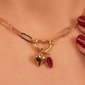 May include: Gold necklace with a heart-shaped pendant, a small heart charm, and a ruby-coloured teardrop charm. The chain is a link design.