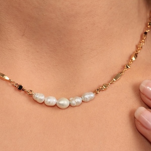 Puede incluir: Un collar dorado con una cadena fina y una fila de cinco perlas blancas naturales. La cadena tiene pequeños eslabones rectangulares. El collar se muestra sobre un fondo color piel.