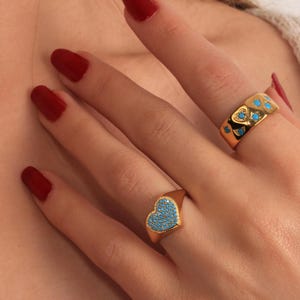 May include: Two gold rings with turquoise detailing. One ring has a heart-shaped face, filled with turquoise stones. The other is a band with heart cutouts and turquoise stones. The rings are on a hand with red nail varnish.