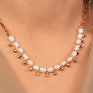 May include: A gold-toned necklace featuring a strand of white, irregularly shaped pearls. Small, gold-colored teardrop-shaped charms are evenly spaced between the pearls. The necklace has a delicate chain and clasp.