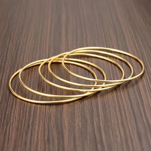 May include: A set of five thin, circular gold-toned bangles arranged in an overlapping, layered formation. Each bangle features a smooth, polished, and continuous metal band with a simple, minimalist design and a reflective, bright yellow-gold finish.