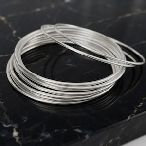 Può includere: Un set di molteplici bracciali rigidi sottili e circolari in argento impilati insieme. I bracciali presentano una finitura metallica liscia e lucida con un design semplice a filo tondo. Ogni bracciale ha uno spessore uniforme.