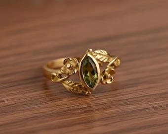 May include: A gold ring featuring a marquise-cut, olive-green gemstone. The ring is adorned with floral and leaf details, creating a nature-inspired design. The ring is set on a wooden surface.