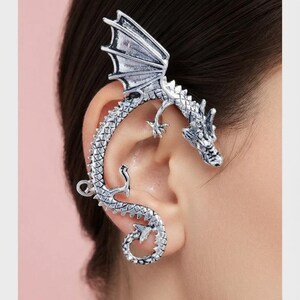 May include: A silver-toned ear cuff shaped like a dragon that wraps around the outer edge of the ear. The dragon features detailed scales, a prominent wing extending upward, and a curled tail at the bottom, with a metallic finish throughout.