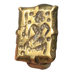 May include: A gold-colored, rectangular pendant with an embossed design. The pendant features a central figure with curved lines and small circular details. The edges are slightly uneven, giving it an antique appearance.