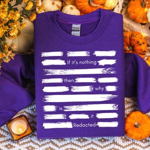 Può includere: Una felpa viola con testo bianco che recita "If it's nothing then why is it redacted". La felpa è circondata da zucche, fiori e una candela accesa.