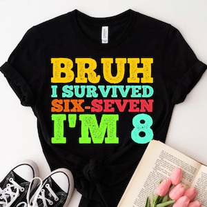 Può includere: T-shirt nero con la scritta "BRUH I SURVIVED SIX-SEVEN I'M 8" in giallo, verde acqua, arancione, rosso e verde. La maglietta è abbinata a scarpe da ginnastica bianche e nere e a un libro aperto con fiori rosa.