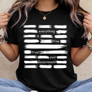 Puede incluir: Camiseta negra con texto blanco y diseño de pinceladas horizontales. El texto dice "everything is fine" y "trust your Government". La camiseta es de cuello redondo y tiene mangas cortas.