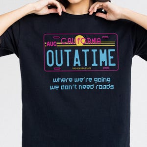 Może przedstawiać: Czarny t-shirt z neonowym wzorem tablicy rejestracyjnej. Projekt zawiera słowo "OUTATIME" oraz tekst "where we're going we don't need roads." Projekt zawiera słowo "California" i żółte słońce.